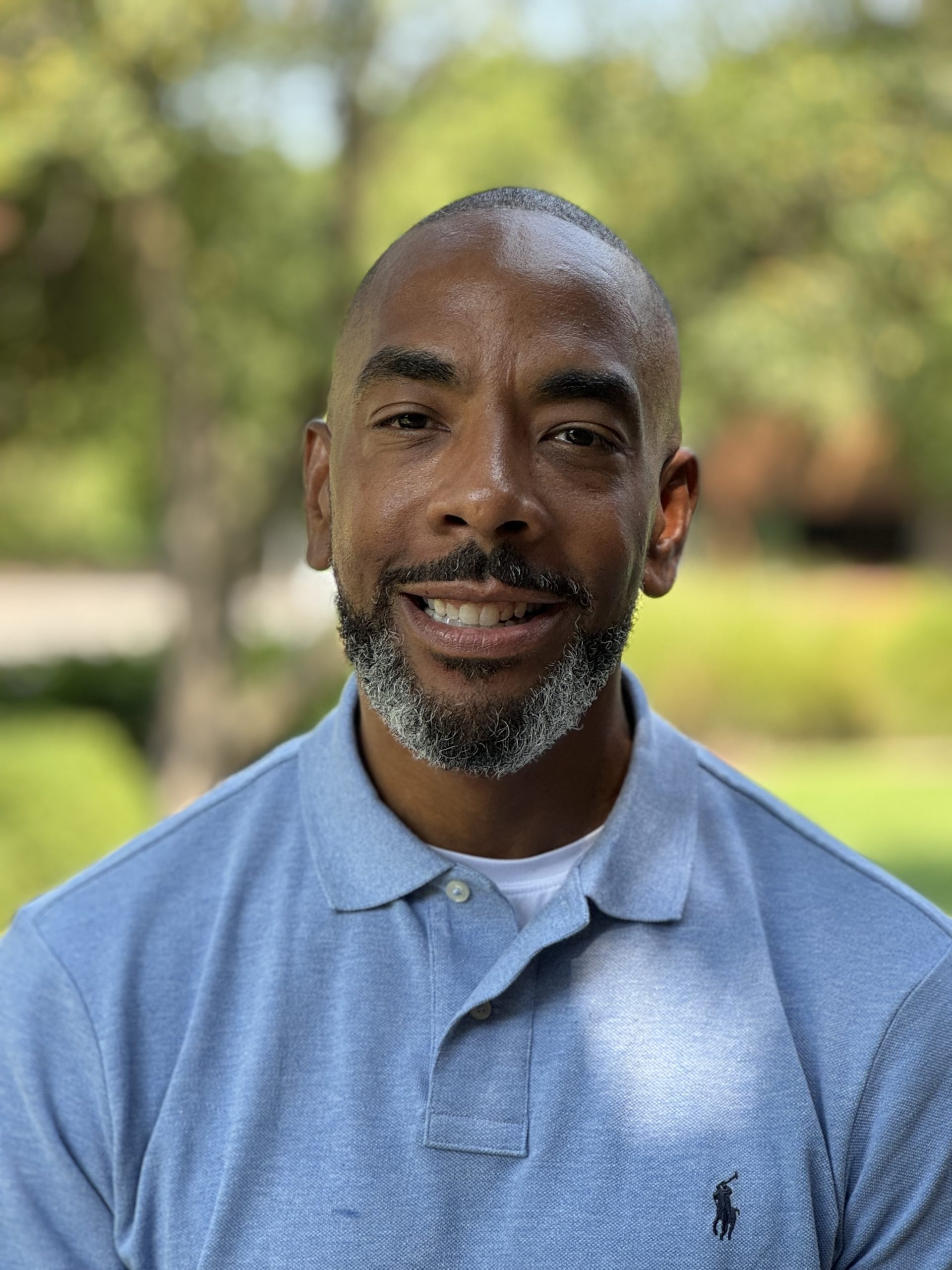 A portrait of Miles Jones wearing blue shirt with blurred background of green trees